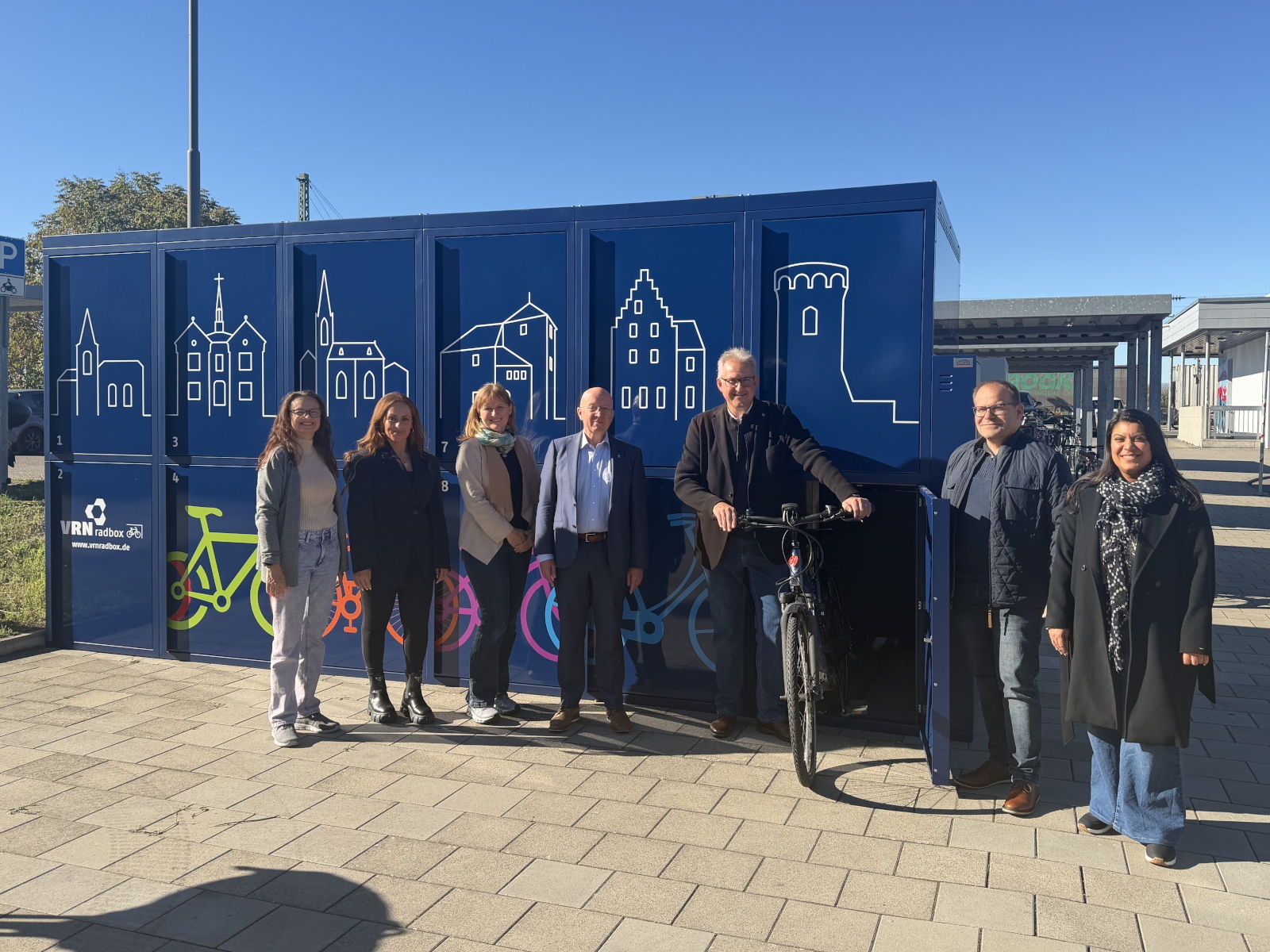 Gruppenfoto vor den blau lackierten Fahrradboxen, auf denen mit weißer Farbe die Silhouetten einiger Gebäude abgebildet sind.